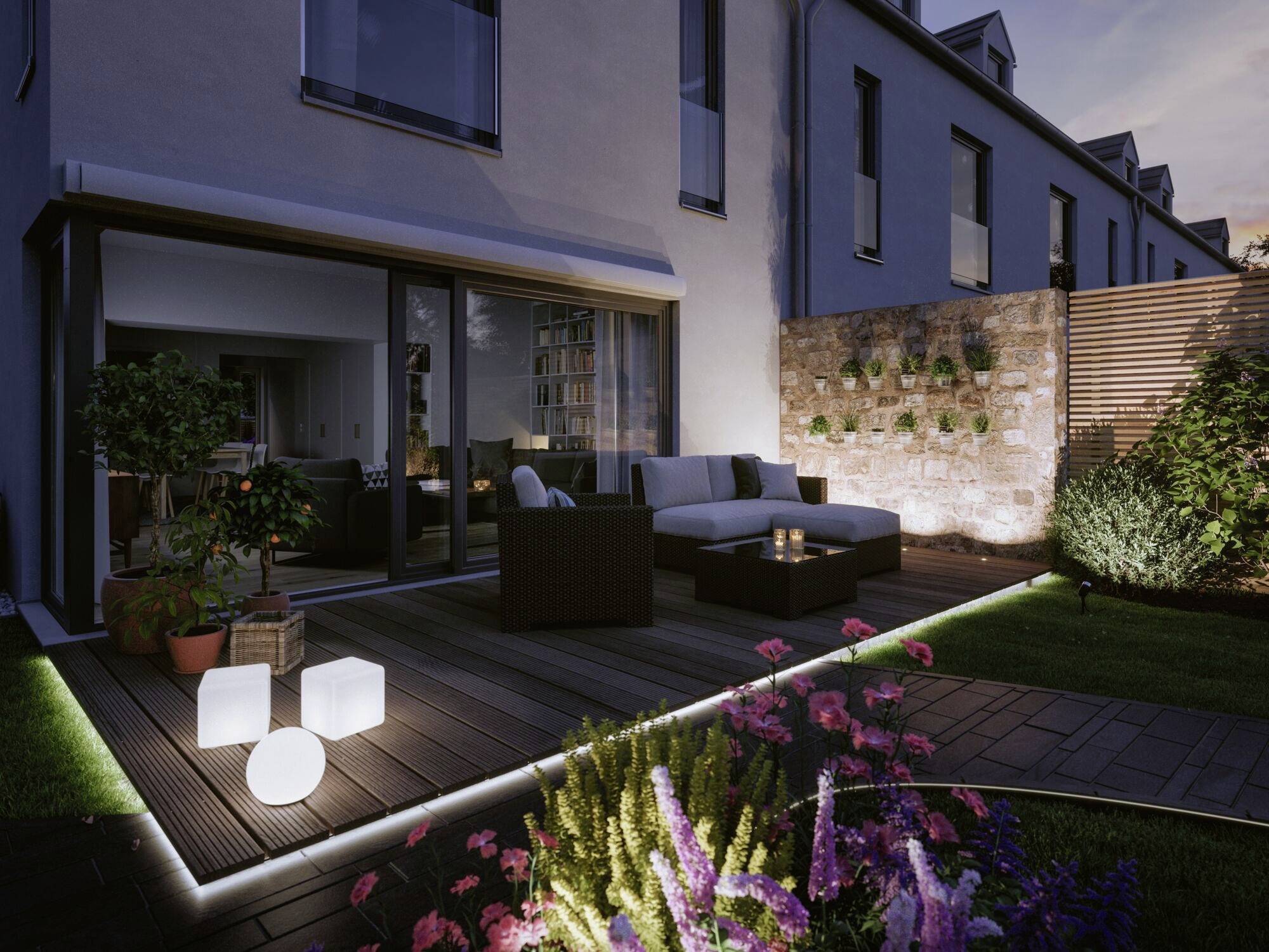 Ein moderner Garten bei Dämmerung mit stilvoll beleuchteter Terrasse, gemütlichen Sitzmöbeln, Pflanzen und vertikalem Kräutergarten.