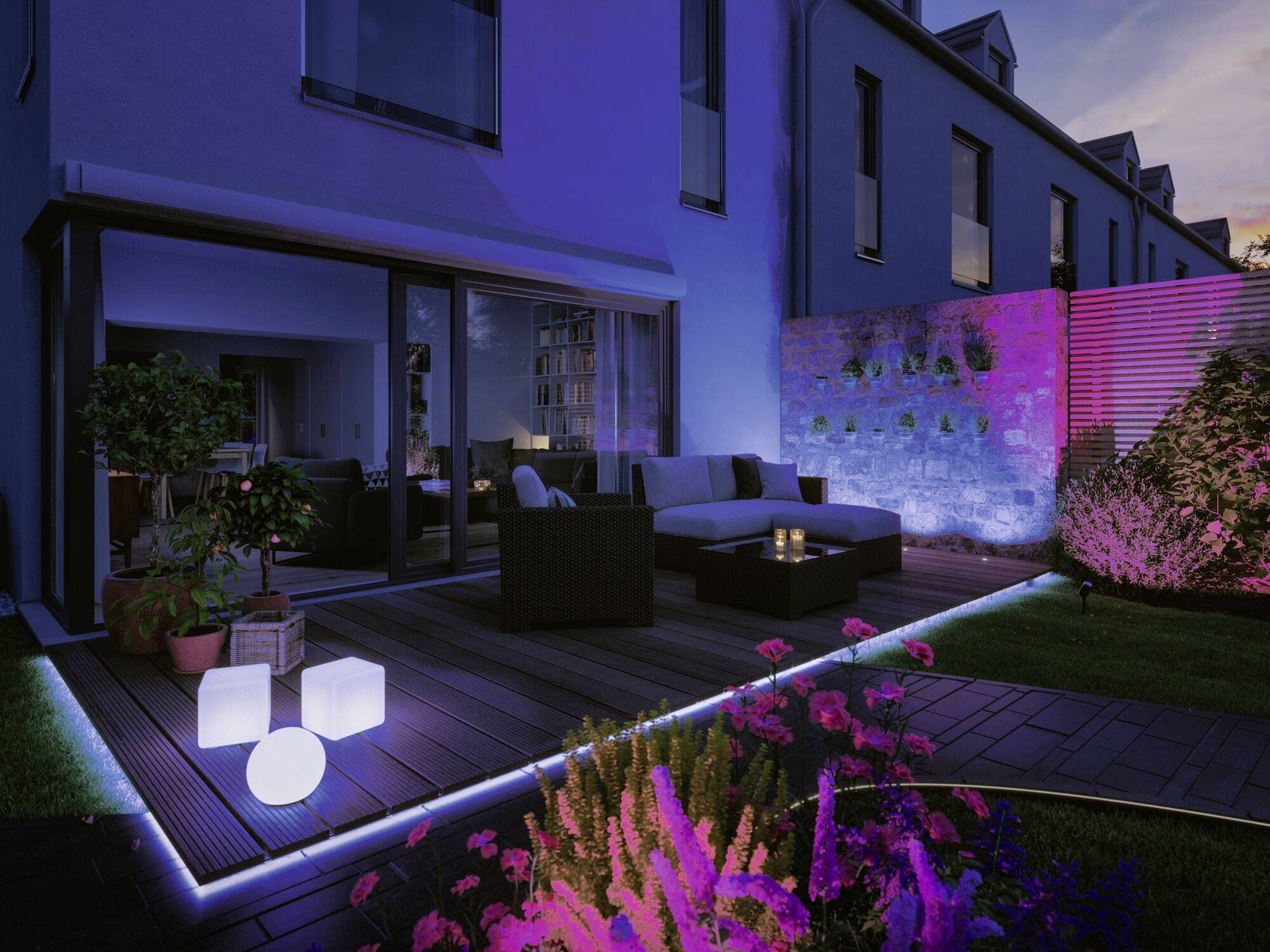 Beleuchtete Terrasse bei Dämmerung mit modernen Gartenmöbeln, farbigen Lichtern und Pflanzen, die eine gemütliche Atmosphäre schaffen.