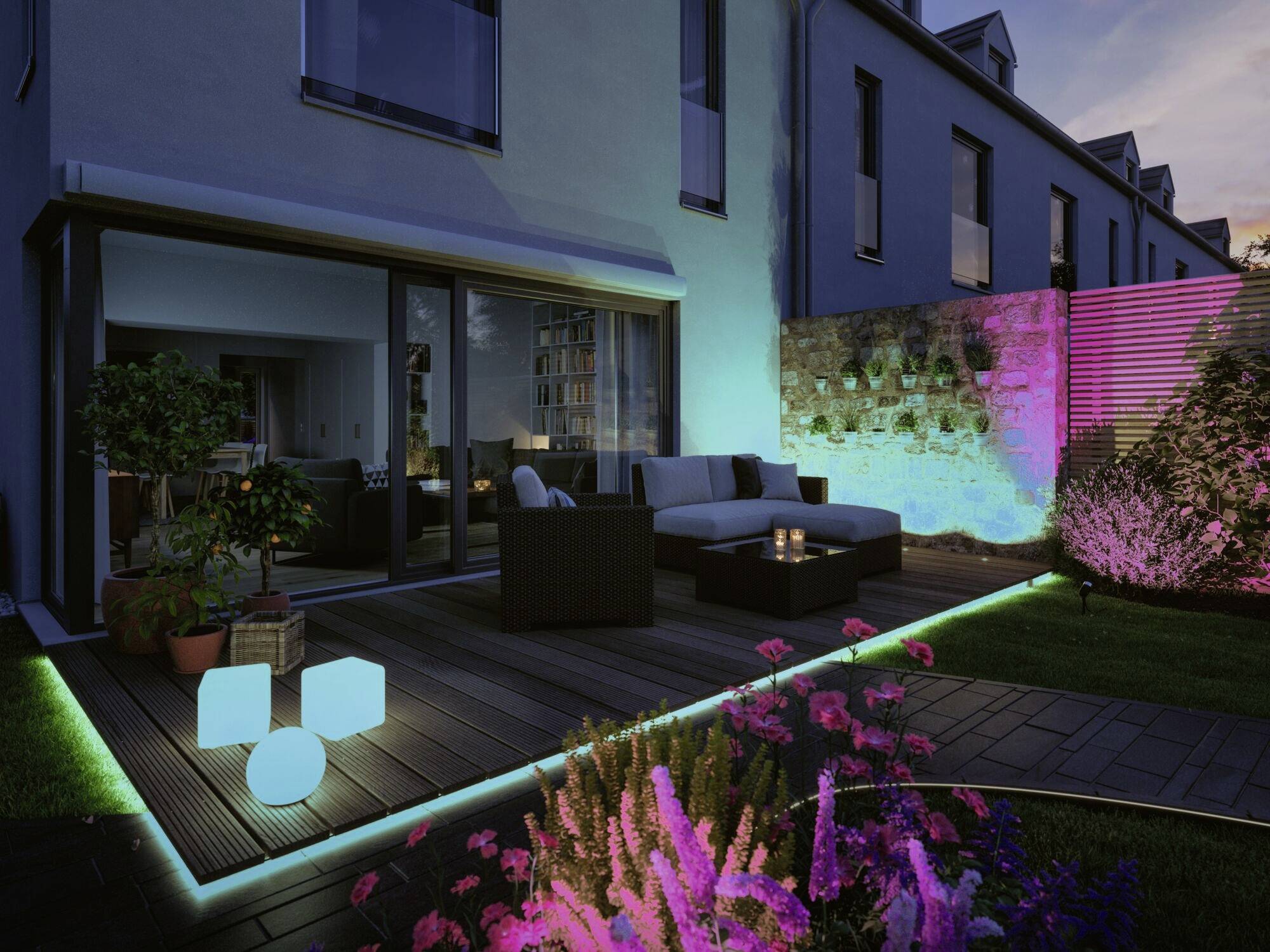 Moderne Terrasse bei Dämmerung, beleuchtet mit farbigen LED-Lichtern. Gartenmöbel und Pflanzen im Vordergrund, Gebäude im Hintergrund.
