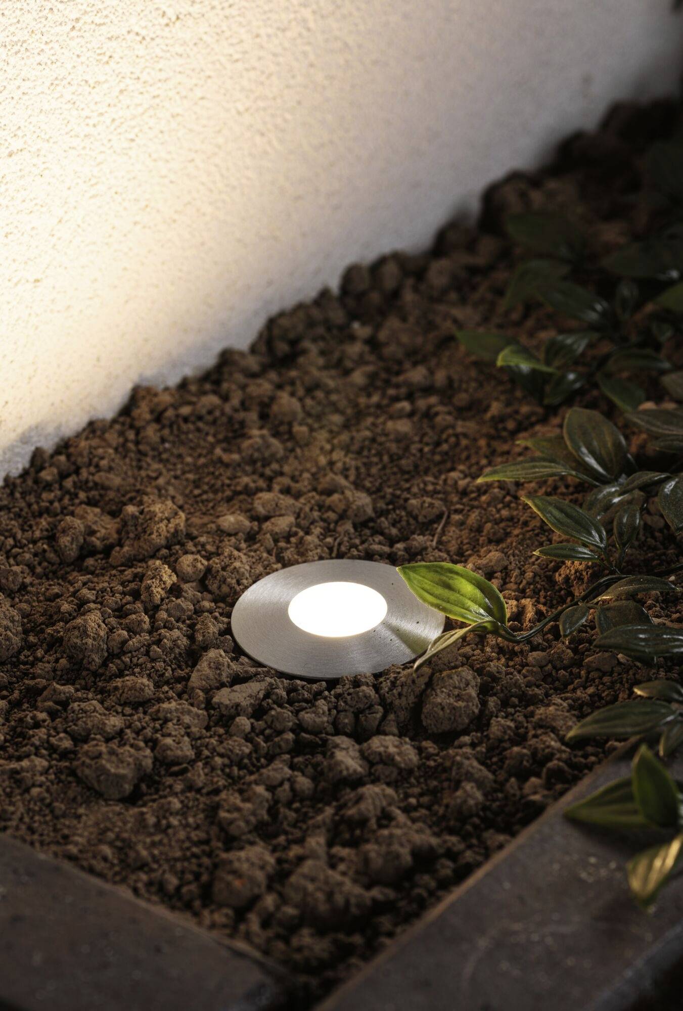 Gartenstrahler im Boden, leuchtet neben einer Wand und Pflanzen. Beleuchtet den Bereich in einem Garten oder Hof für Ambiente oder Sicherheit.