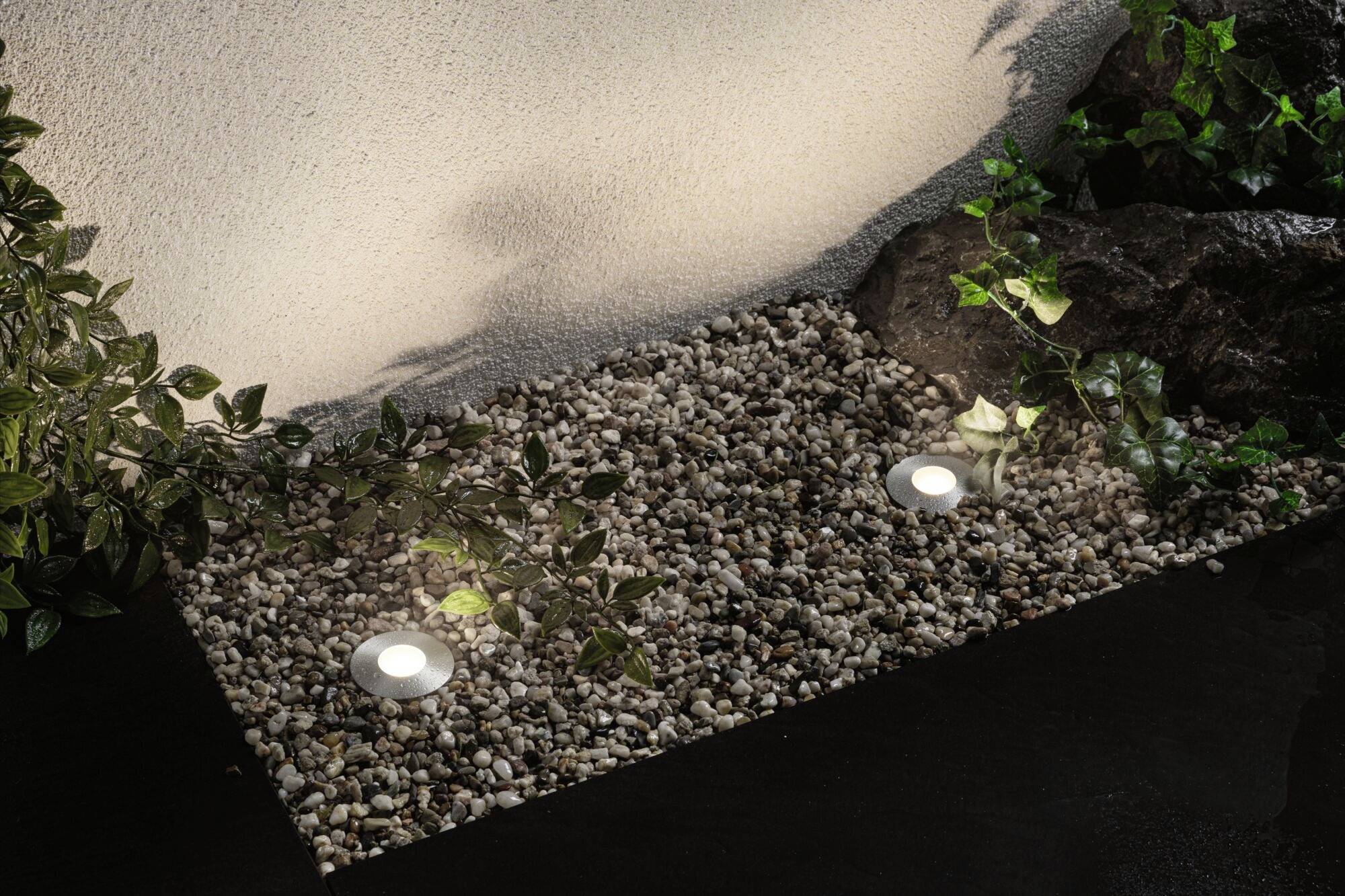 Gartenweg mit kleinen Kieselsteinen und zwei runden Bodenlichtern, umgeben von grünen Pflanzen und einem Felsen am Rand.