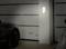 Garage mit zwei Autos, schwach beleuchtet durch eine Wandlampe. Rahmen der Garagentür im Hintergrund sichtbar.