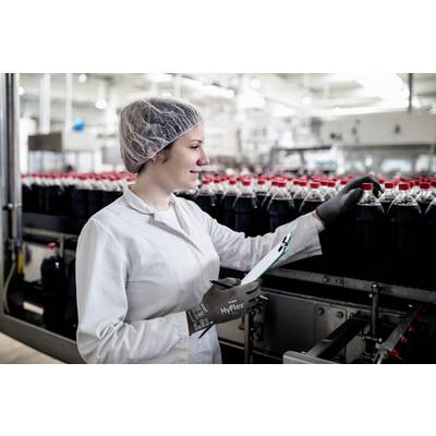 Eine Person in weißem Kittel und Haarnetz inspiziert Flaschen an einem Fließband in einer Fabrik. Sie hält ein Klemmbrett.