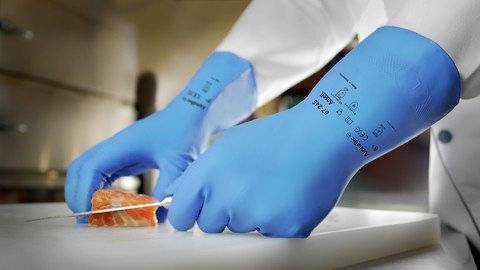 Eine Person mit blauen Handschuhen schneidet Lachsfilet auf einem Schneidebrett in einer Küchenumgebung.