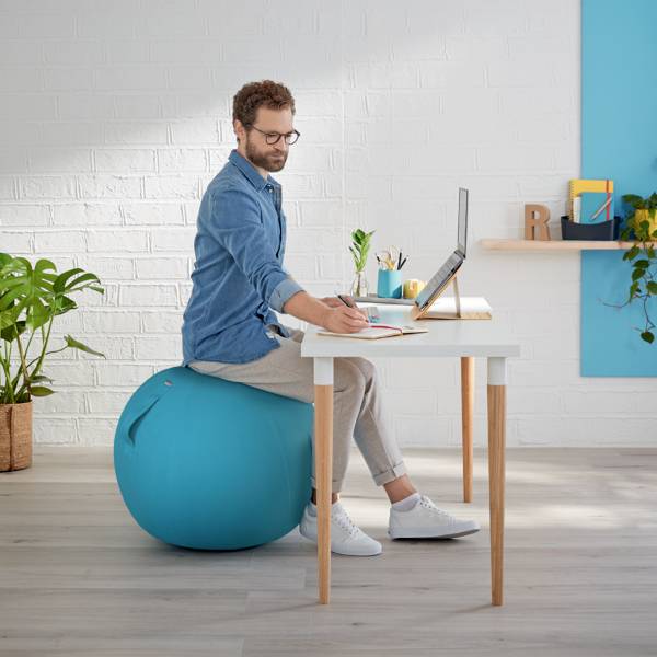 Eine Person sitzt an einem Schreibtisch, schreibt in ein Notizbuch und nutzt einen Laptop. Eine Pflanze und Dekoration sind im Hintergrund.