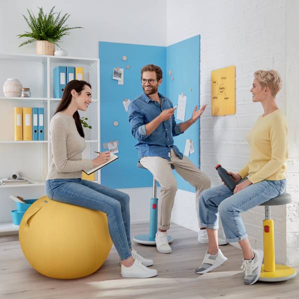 Drei Personen in einem modernen Büro, sitzen auf ergonomischen Stühlen und einem Ball. Sie diskutieren an einer Wand mit Notizen.