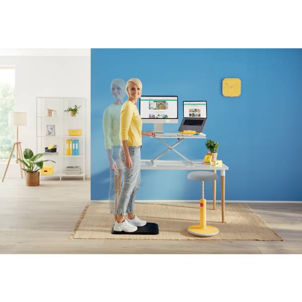 Eine Person in Freizeitkleidung steht auf einem Balance Board vor einem höhenverstellbaren Schreibtisch mit zwei Monitoren, einer Pflanze und moderner Einrichtung im Hintergrund.