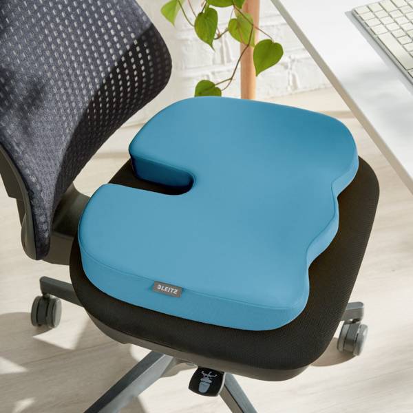 Ein ergonomisches Sitzkissen auf einem Bürostuhl, designed für verbesserten Sitzkomfort. Daneben ein Tisch mit einer Pflanze im Hintergrund.