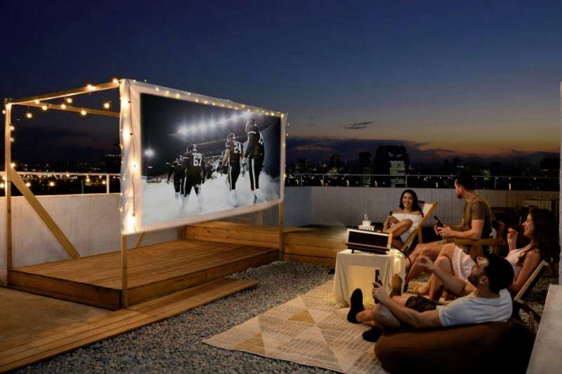 Personen sitzen auf einer Dachterrasse und schauen einen Film auf einer Leinwand bei Sonnenuntergang. Lichterketten dekorieren die Leinwand.