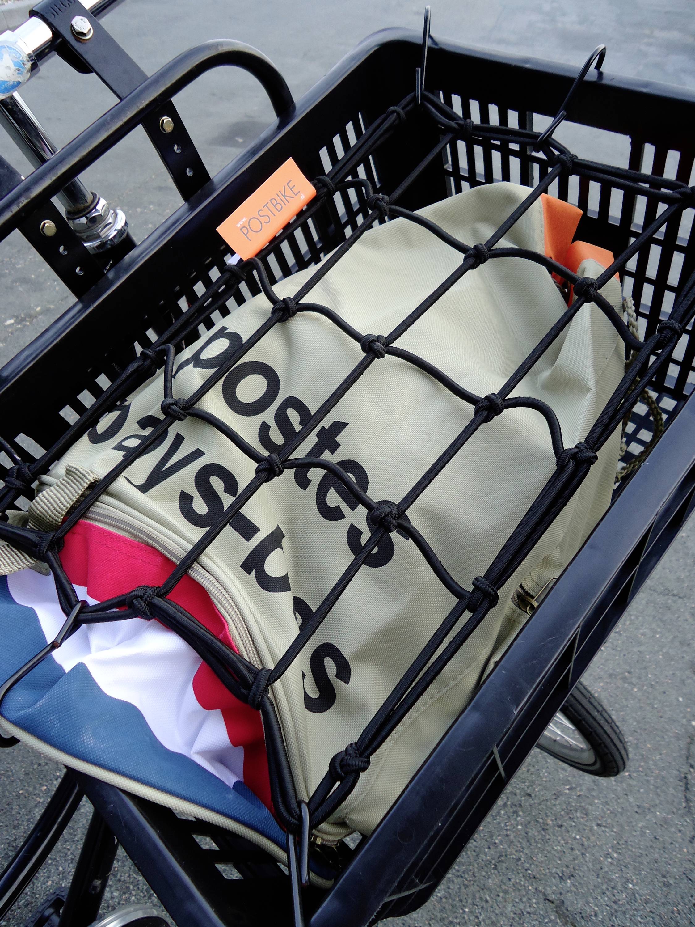 Ein Fahrradkorb enthält eine beigefarbene Tasche mit der Aufschrift 'Booster Bag Sport', gesichert durch ein schwarzes Netz.