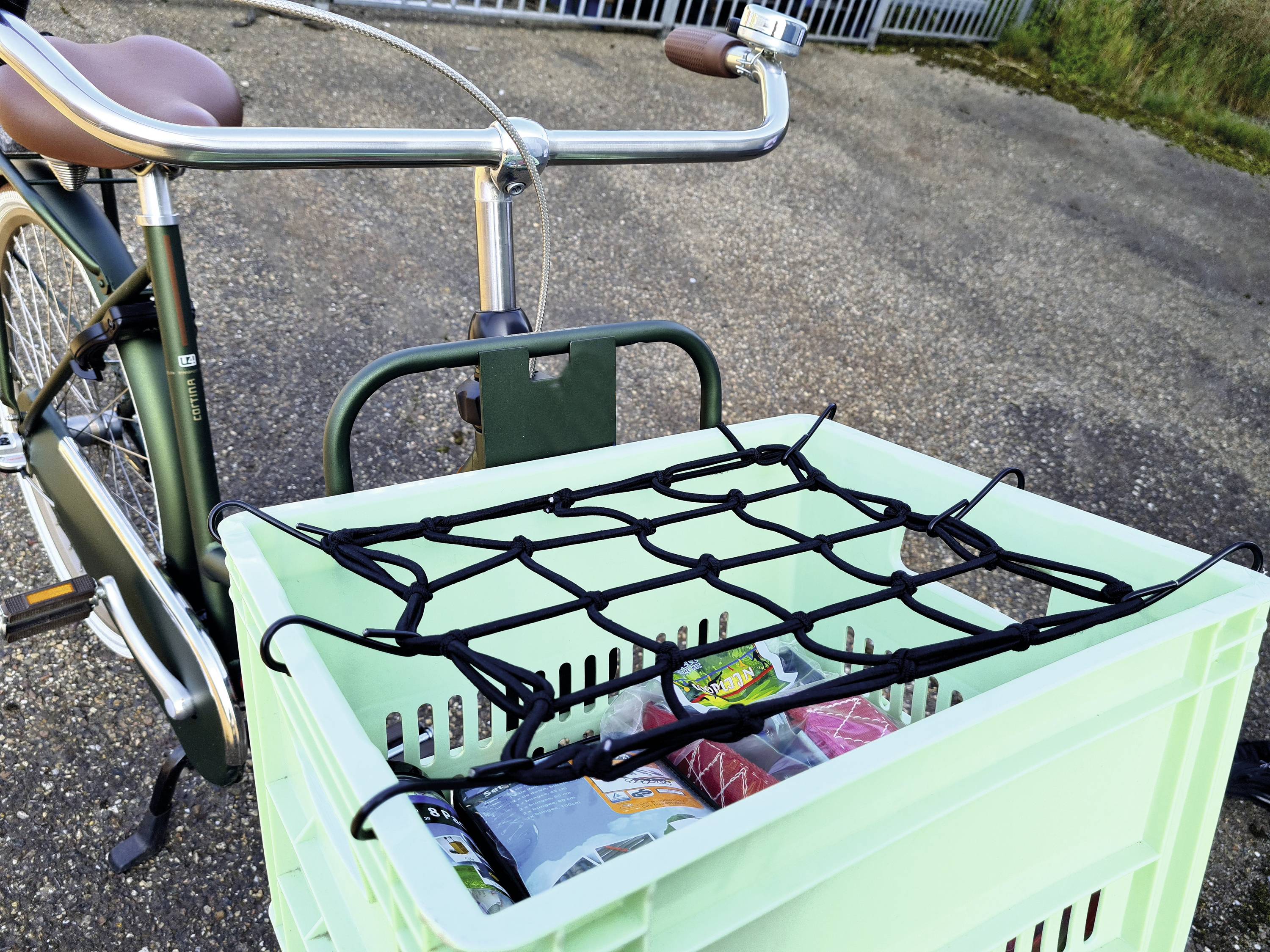 Fahrrad mit grünem Transportkorb hinten, der mit einem Netz gesichert ist. Der Korb enthält Einkaufstüten und steht auf einem Asphaltweg.