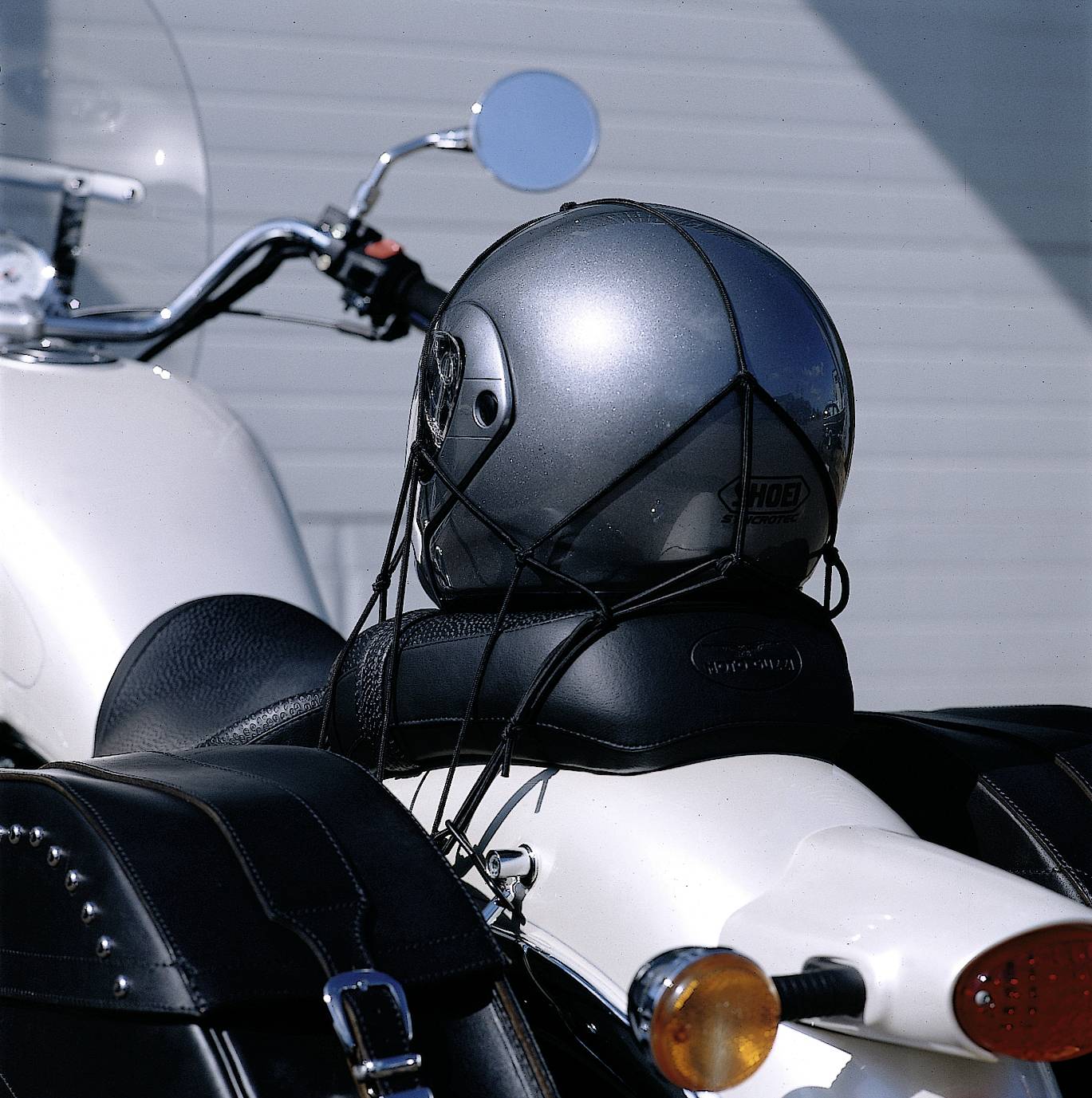 Ein silberner Motorradhelm ist auf dem Sitz eines weißen Motorrads befestigt. Links und rechts sind schwarze Packtaschen sichtbar.