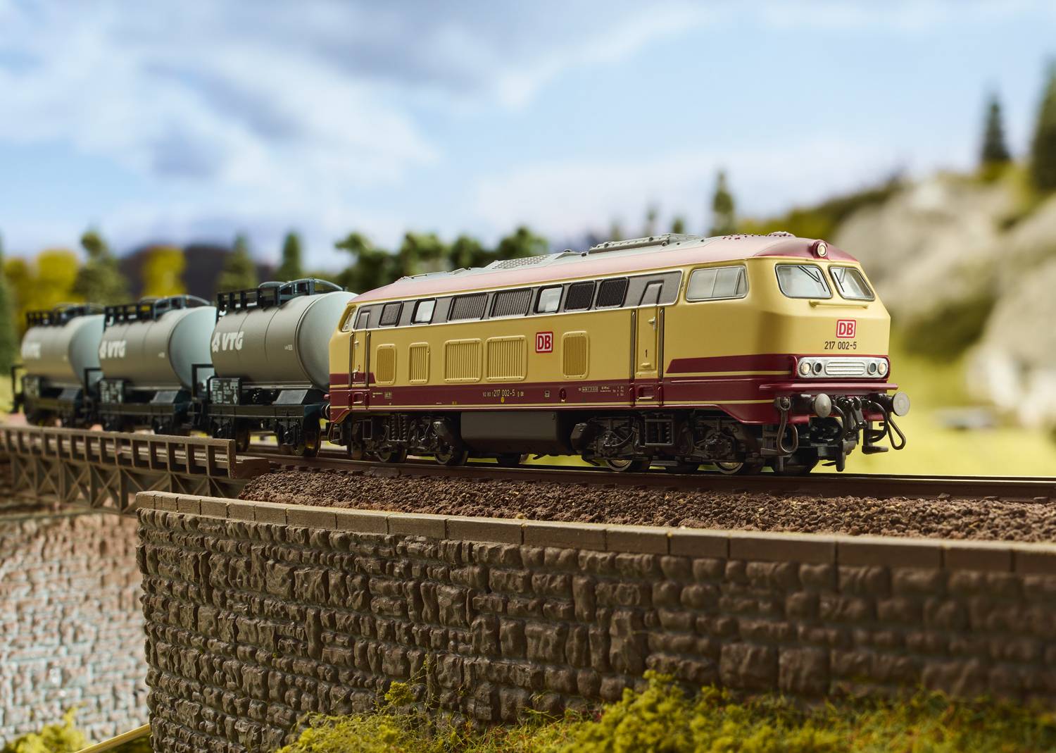 Eine Modelllokomotive mit zwei Kesselwaggons auf einer Brücke in Landschaftsdarstellung. Die Lok ist in rot-beiger DB-Lackierung gehalten.