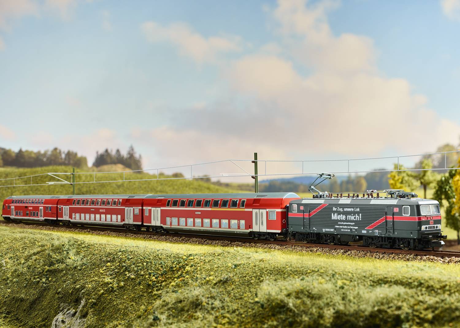 Zug mit roten Doppeldeckerwaggons fährt über eine grüne Landschaft unter blauem Himmel. 