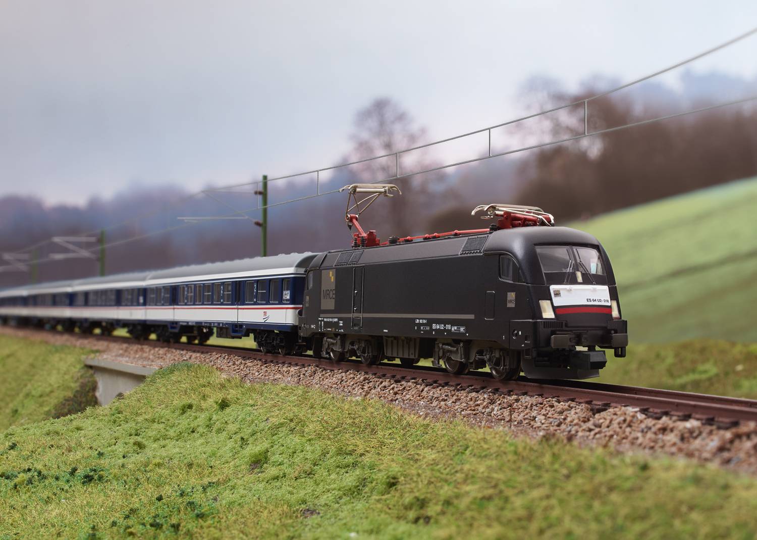 Ein schwarz-weißer Zug fährt mit mehreren Waggons über eine Schienenstrecke in einer ländlichen Landschaft mit grünen Wiesen und Bäumen.