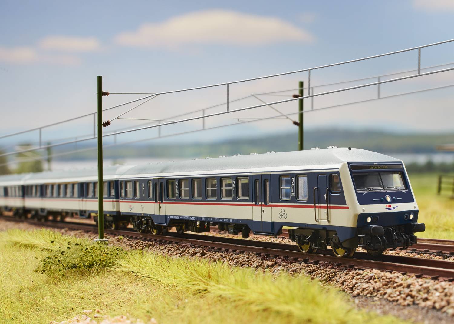 Ein moderner Zug fährt bei sonnigem Wetter über eine ländliche Eisenbahnstrecke. Im Hintergrund sind Stromleitungen und Wiesen zu sehen.