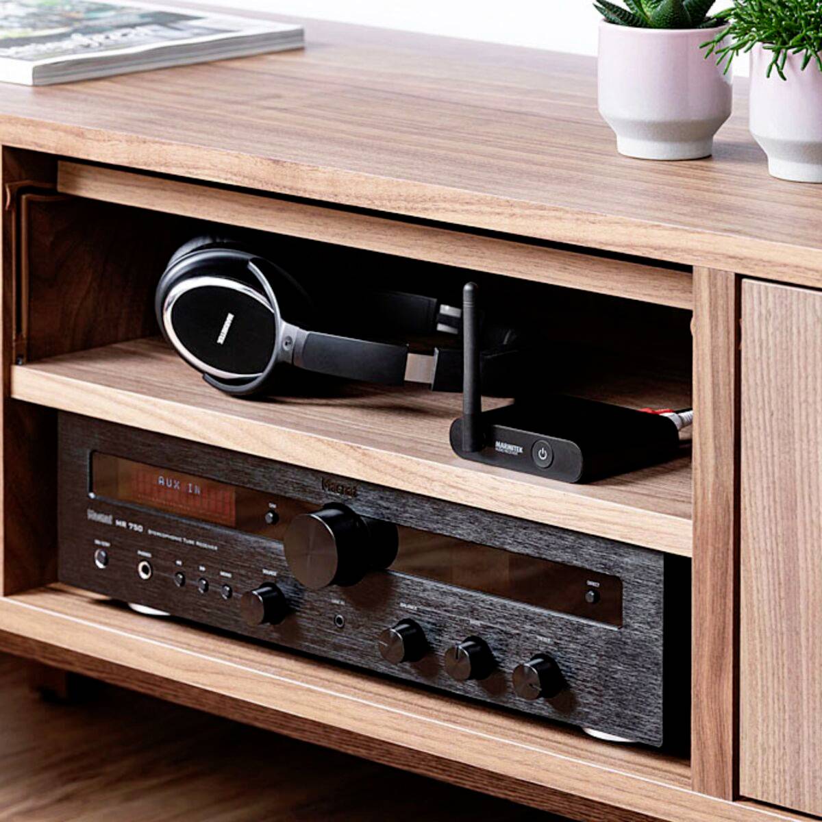 In einem Holzregal stehen Kopfhörer, ein WLAN-Router und ein Audioverstärker. Oben liegt eine Zeitschrift, daneben eine Pflanze.