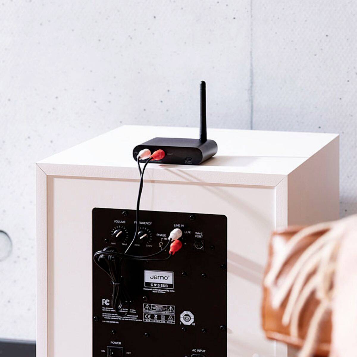 Ein schwarzer drahtloser Audio-Adapter mit einer Antenne und zwei eingesteckten roten Kabeln steht auf einem weißen Lautsprecher.