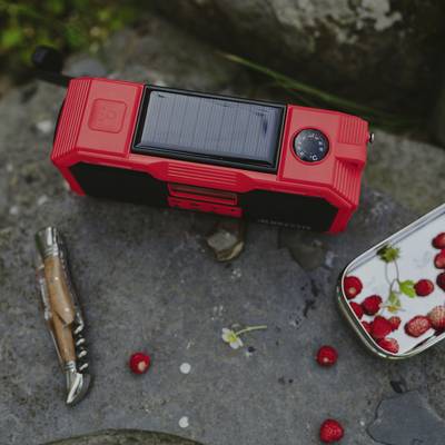 Ein rotes tragbares Radiogerät mit Solarpanel liegt auf einem Stein, umgeben von Himbeeren und einem Taschenmesser.