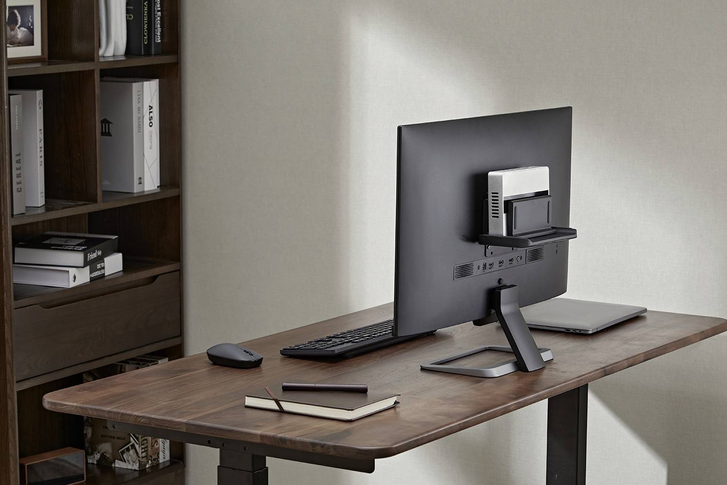 Ein moderner Schreibtisch mit einem Monitor, einer Tastatur, einem Laptop, zwei Notizbüchern und einer Maus in einem Büro mit Bücherregal.