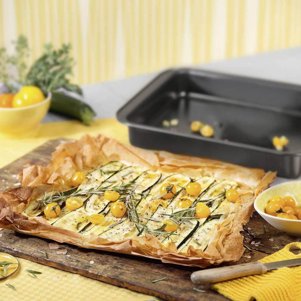 Quiche mit Zucchini und gelben Tomaten auf einem Holzbrett, frisch gebacken. Im Hintergrund Kräuter und ein leeres Backblech.