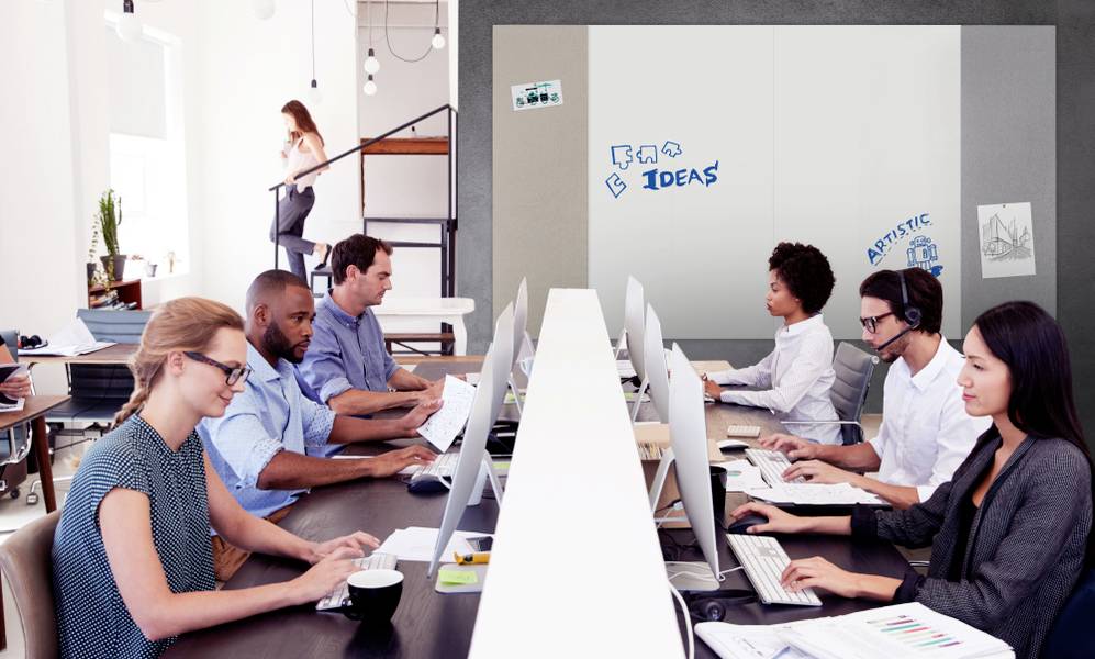 Menschen arbeiten an Computern in einem modernen Büro. Im Hintergrund steht eine Frau auf einer Treppe. An der Wand steht 'Ideas'.