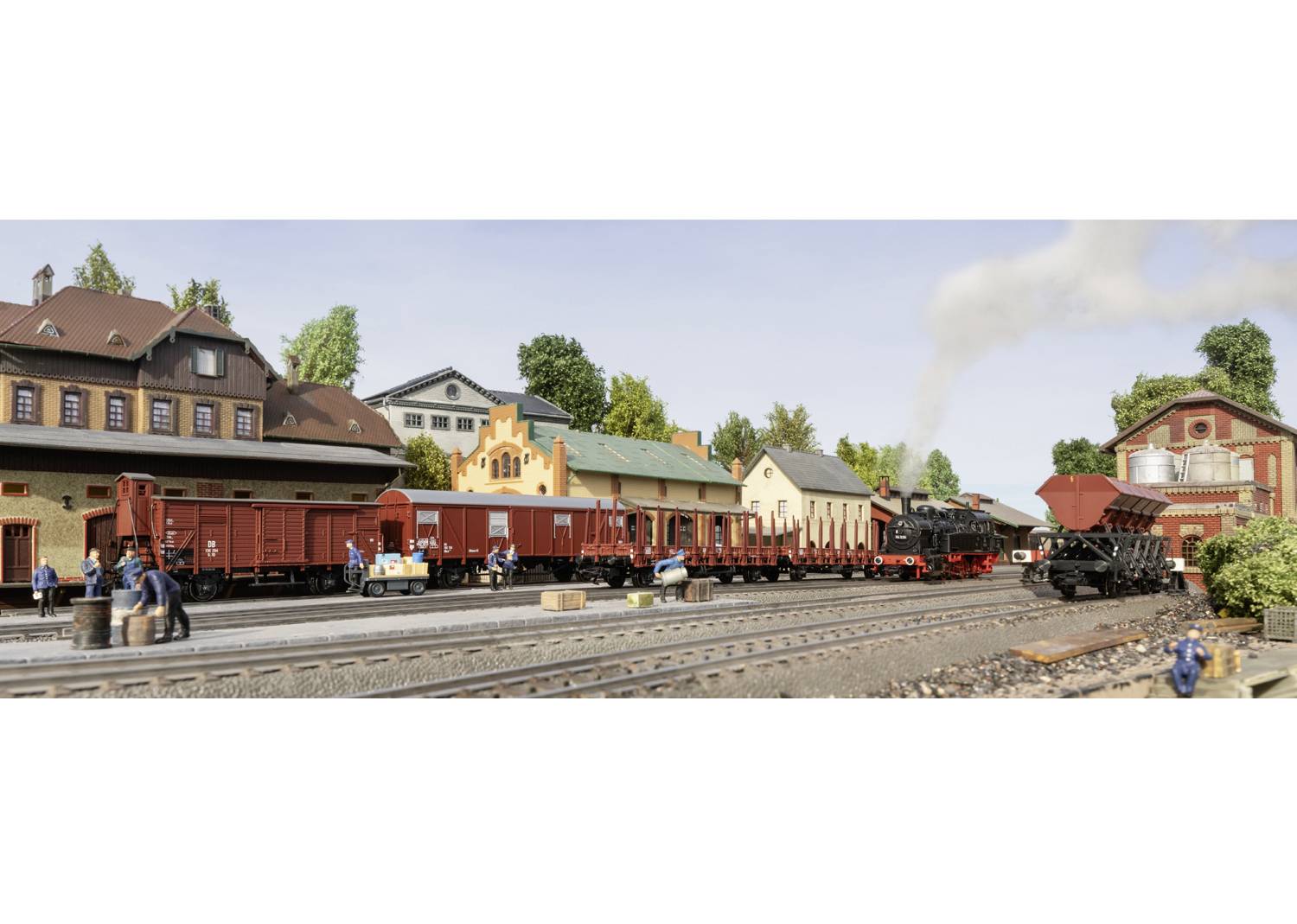 Modelleisenbahnszene mit Güterzug auf Bahnsteig in städtischer Umgebung, inklusive Dampflokomotive und Waggons, umgeben von Gebäuden und Bäumen.