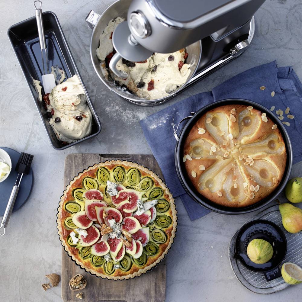 Ein Küchenarbeitsplatz mit einem Mixer voller Teig, zwei fertigen Obsttorten (eine mit Feigen, eine mit Birnen) und Zutaten.