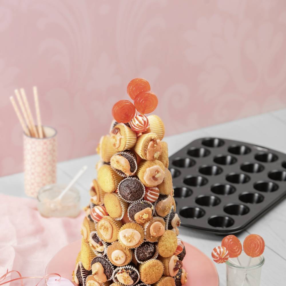 Ein pyramidenförmiger Turm aus Mini-Cupcakes mit dekorativen roten und orangefarbenen Akzenten, auf einem Tisch mit Backzubehör.