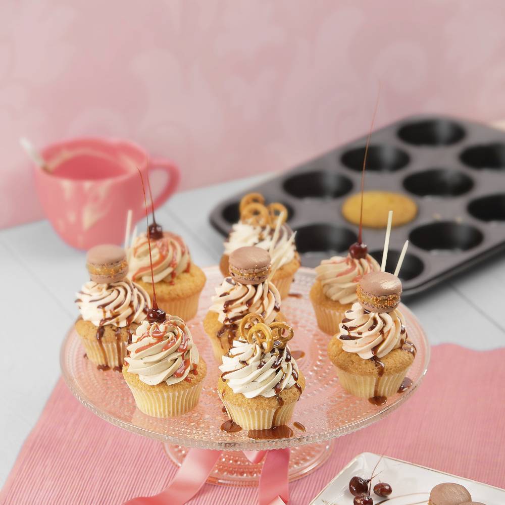 Mehrere kunstvoll verzierte Cupcakes mit Kirschen auf einer Glasplatte; Cupcake-Form im Hintergrund.