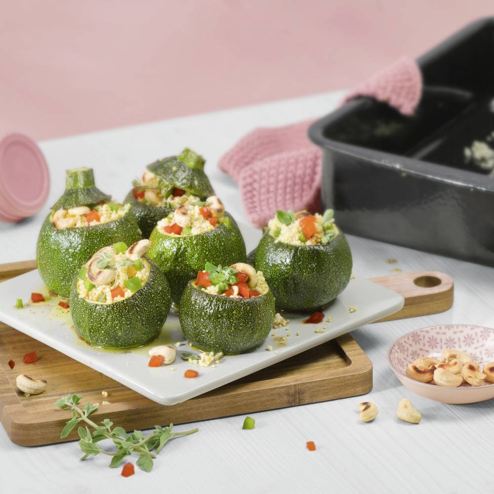 Runde Zucchini auf weißem Teller, gefüllt mit Paprika, Nüssen und Kräutern. Im Hintergrund eine Backform und rosafarbene Stoffserviette.