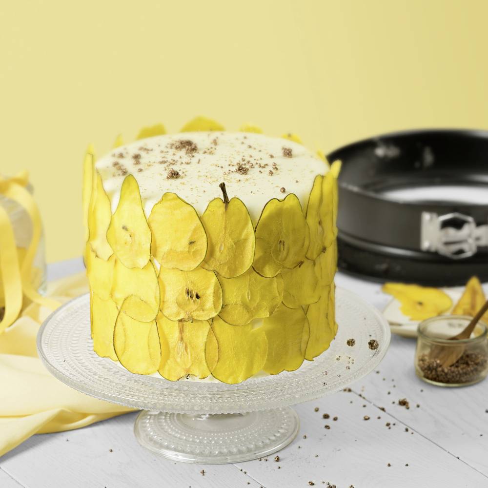 Ein Kuchen mit gelben Apfelscheiben als Dekoration, dekoriert mit weißem Zuckerguss und Schokoladenstückchen auf einem Glasständer.