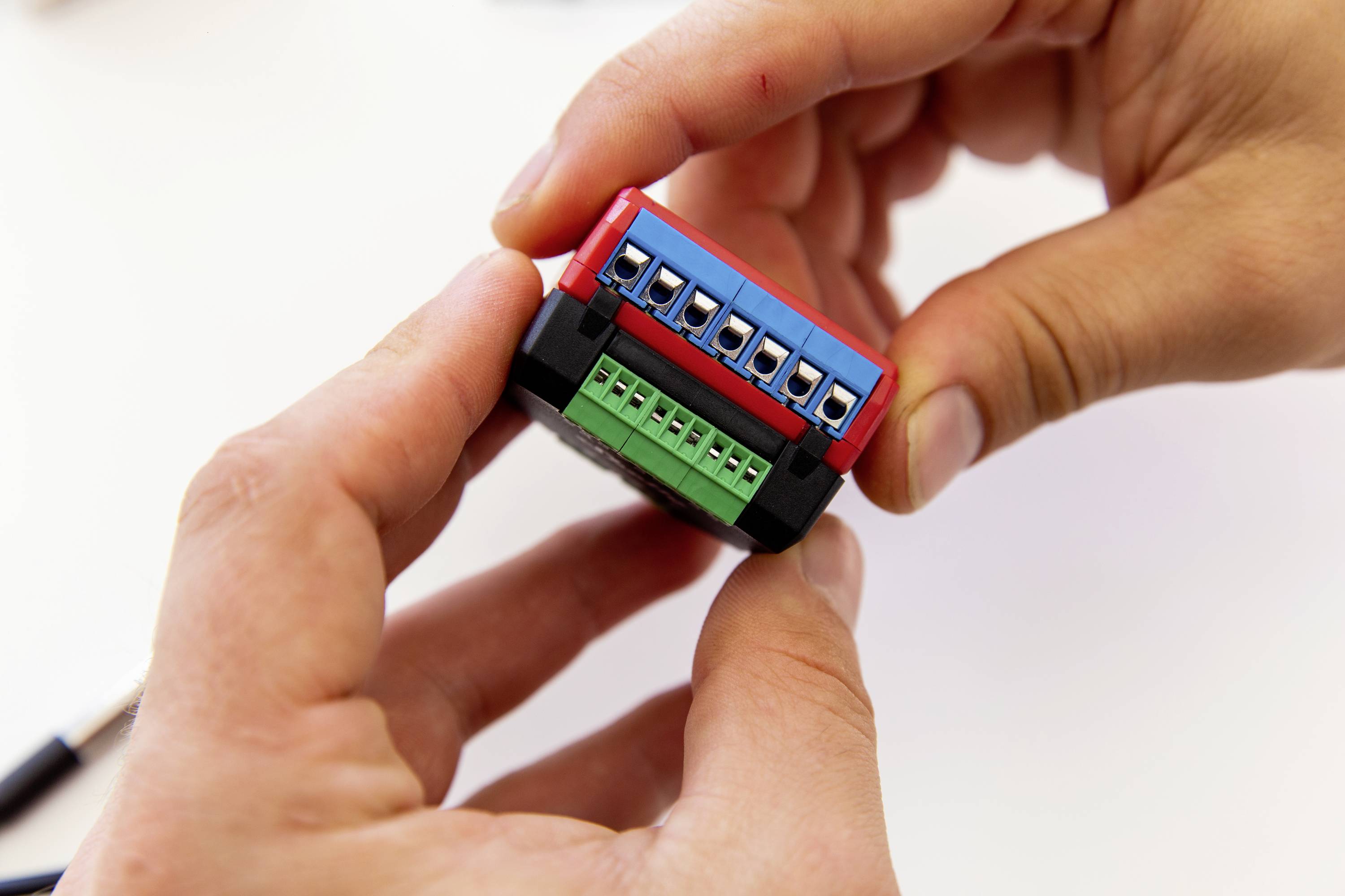 Zwei Hände halten ein kleines elektronisches Gerät mit blauen und grünen Anschlussklemmen, möglicherweise ein Adapter oder Sensor.