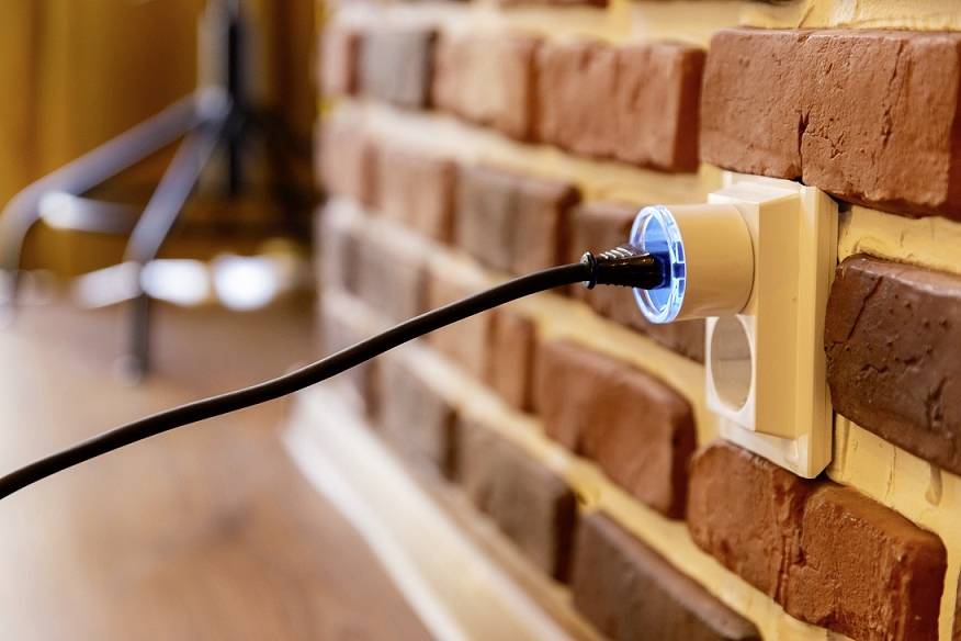 Ein schwarzes Kabel ist in eine Steckdose in einer Ziegelwand eingesteckt. Die Steckdose leuchtet blau und befindet sich im Vordergrund.