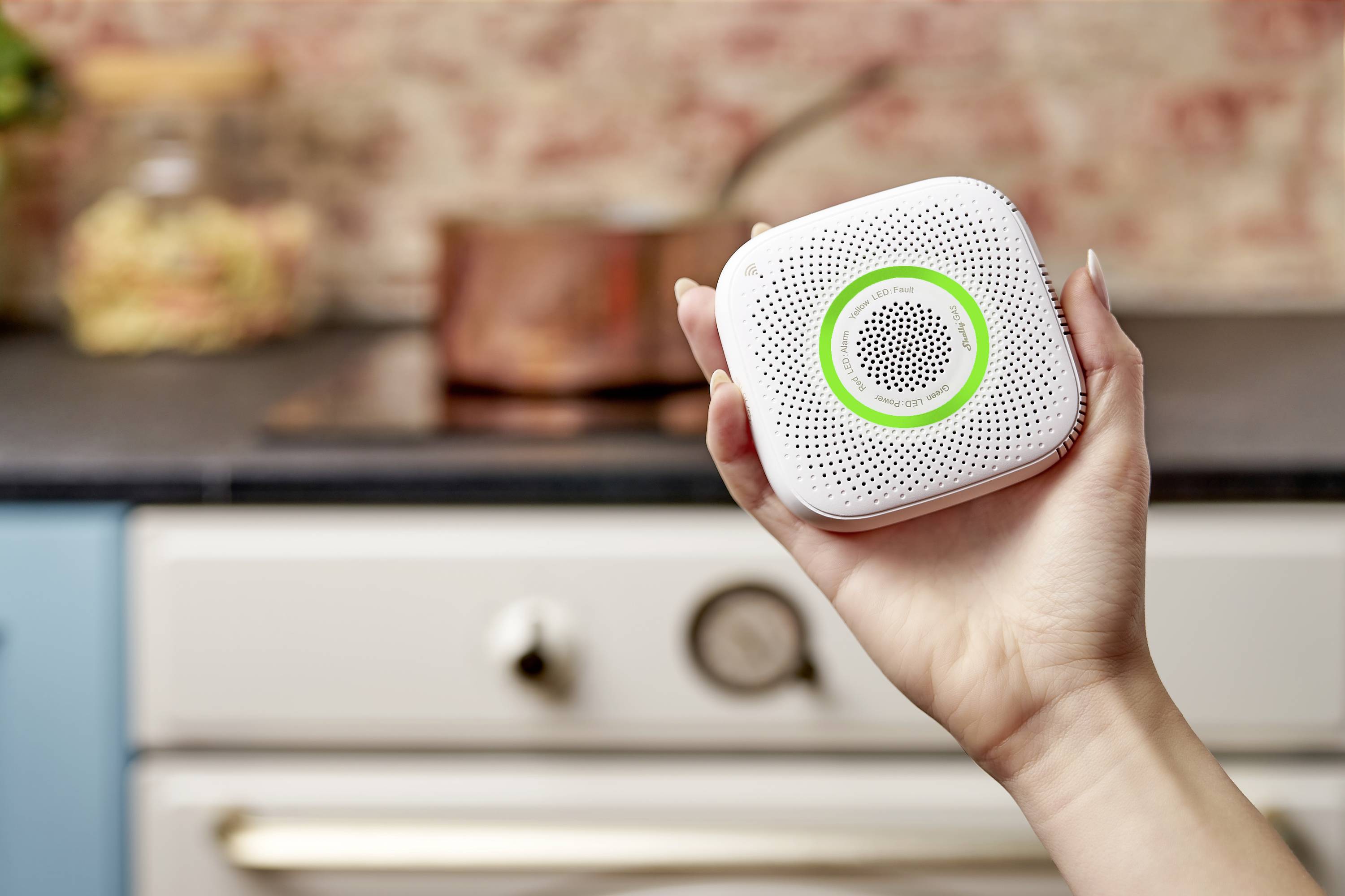 Eine Hand hält einen kleinen, weißen Rauchmelder mit einem grünen Ring, im Hintergrund ist eine Küche mit einem Topf zu sehen.