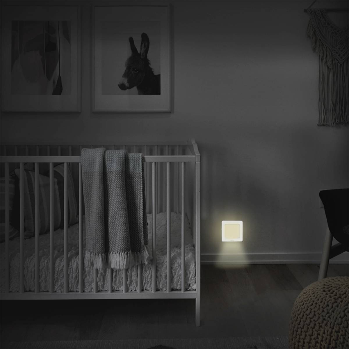 Ein Kinderzimmer bei Nacht mit einem beleuchteten Nachtlicht an der Wand neben einem weißen Kinderbett mit grauer und blauer Decke.