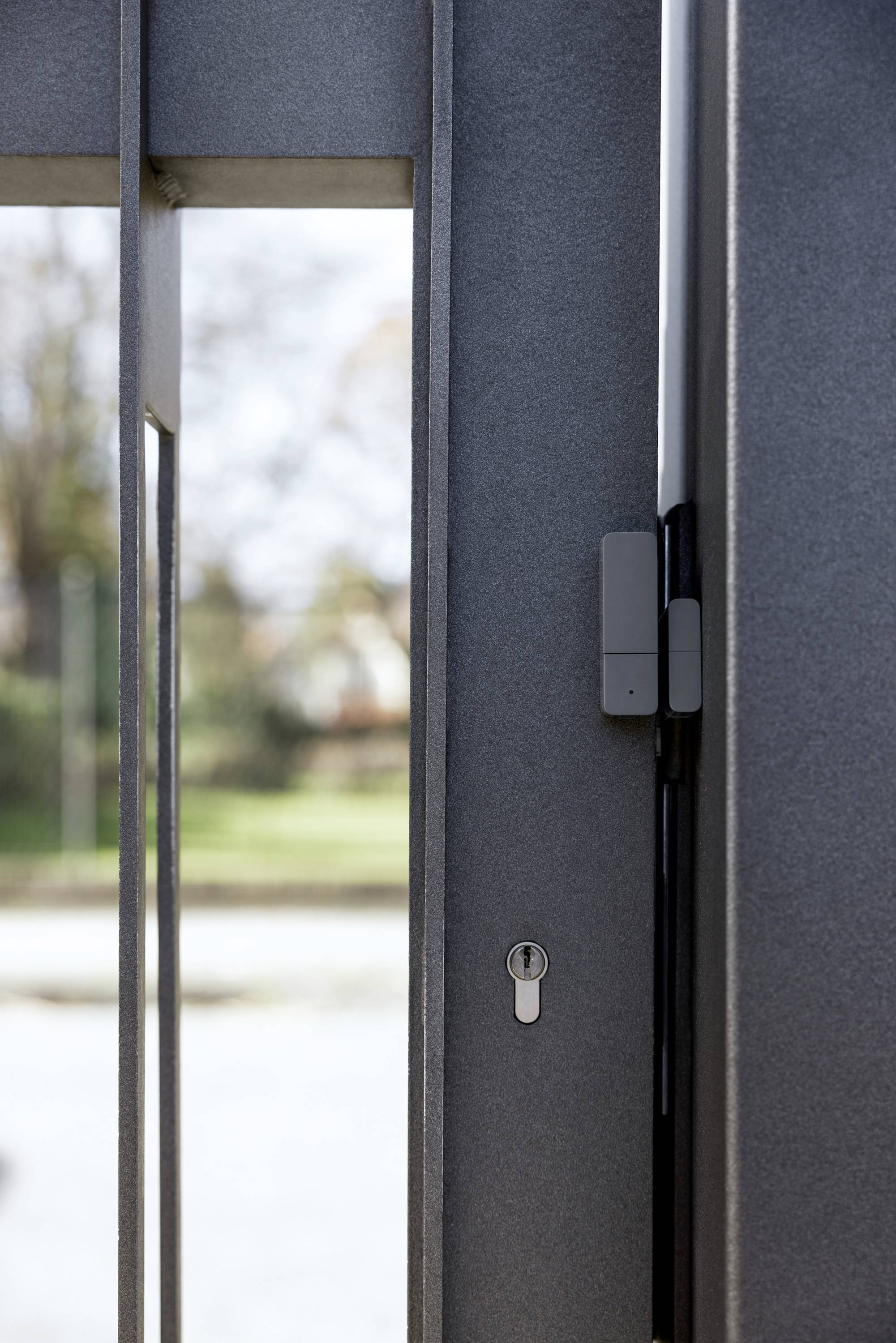 Eine metallene Tür steht offen mit einem Fokus auf das Schloss. Im Hintergrund sind unscharfe Bäume und Rasen zu sehen.