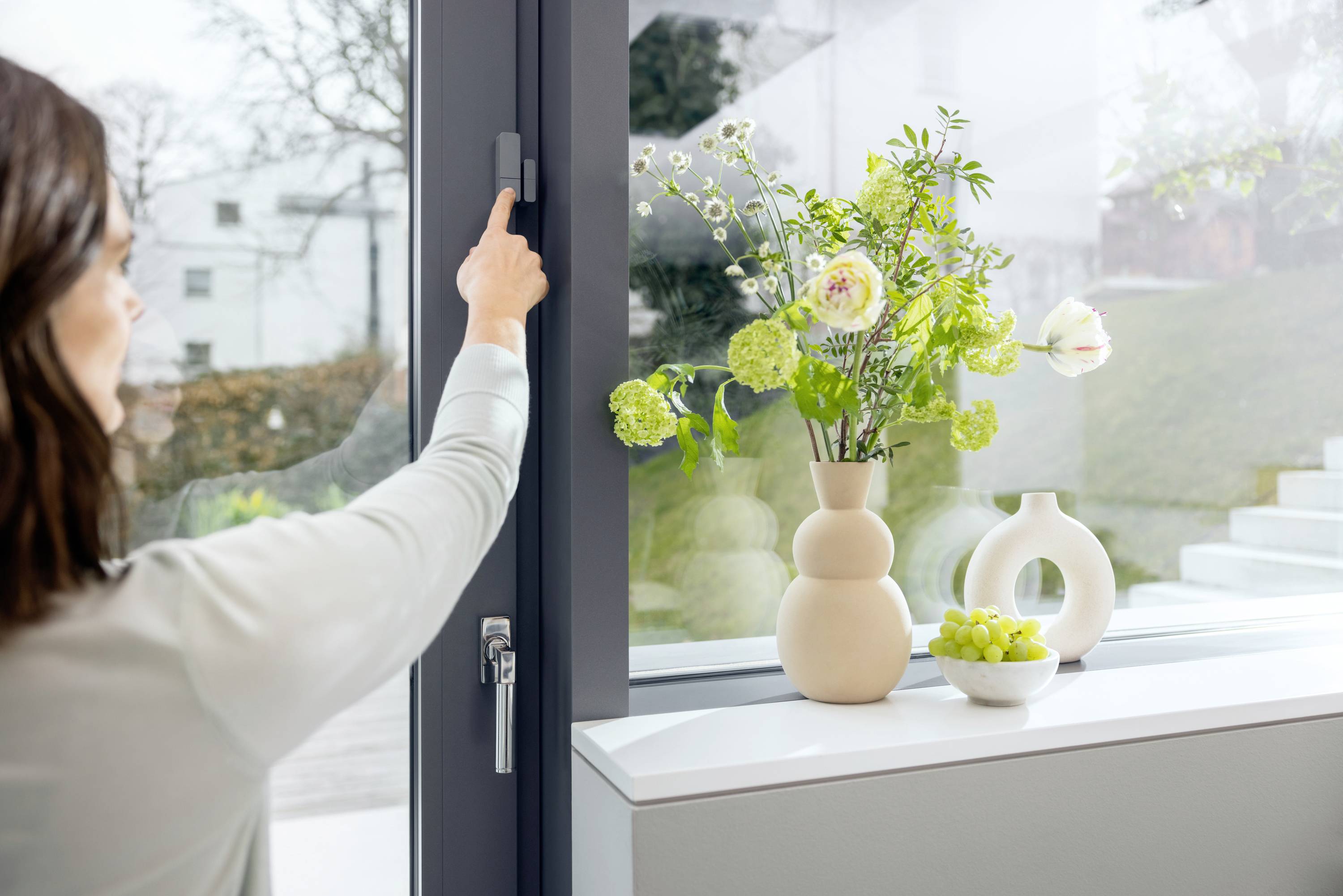 Eine Frau öffnet ein Fenstergriff. Auf der Fensterbank stehen zwei Vasen mit Blumen und eine Schale mit grünen Trauben.
