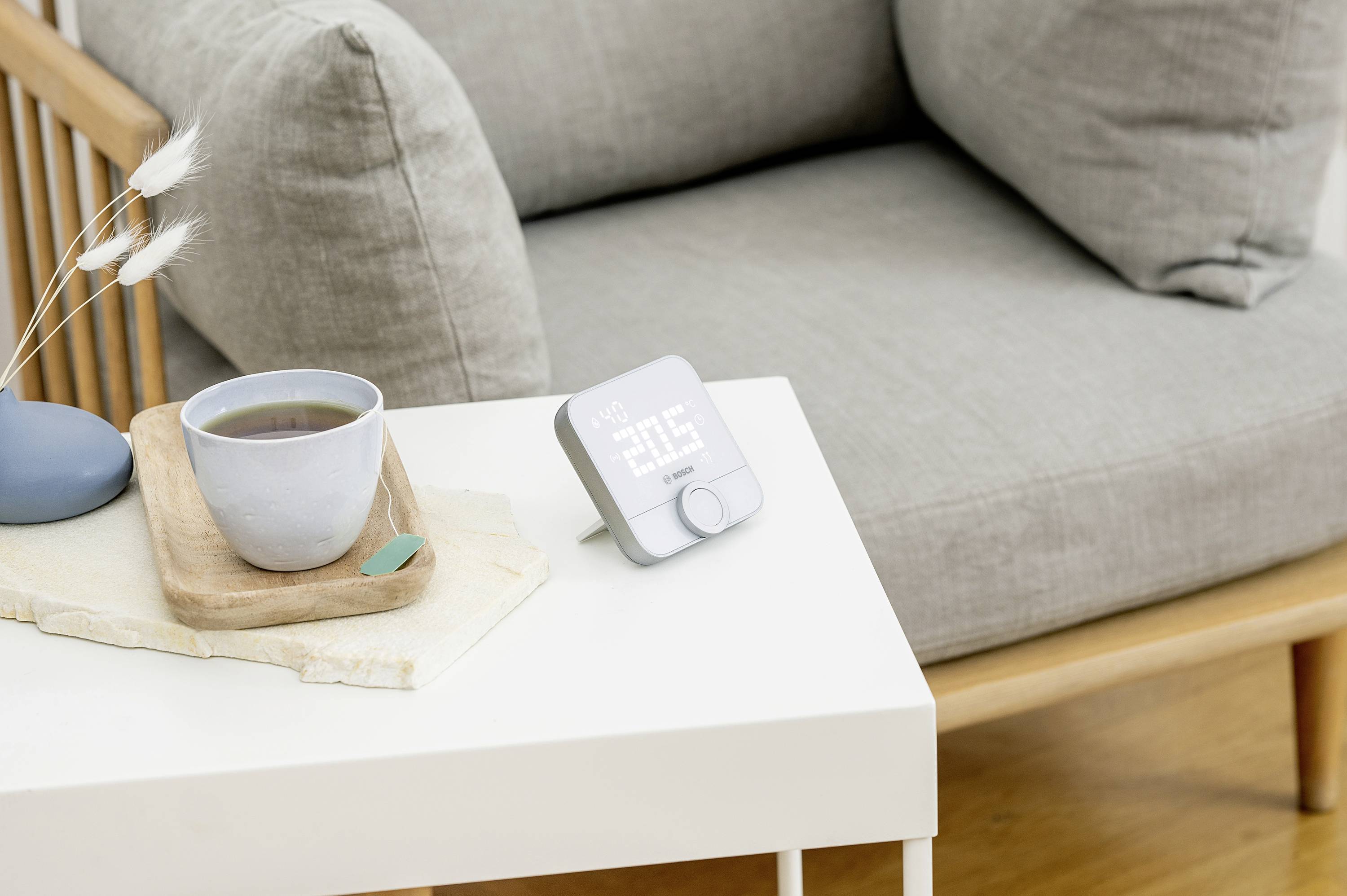 Ein smarter Wecker mit weißem LED-Display zeigt '07:30'. Er steht auf einem weißen Tisch neben einer Tasse Tee nahe einem grauen Sessel.