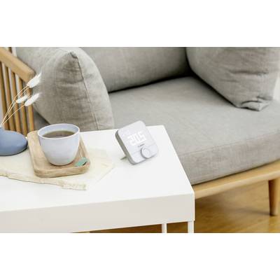 Ein smarter Wecker mit weißem LED-Display zeigt '07:30'. Er steht auf einem weißen Tisch neben einer Tasse Tee nahe einem grauen Sessel.