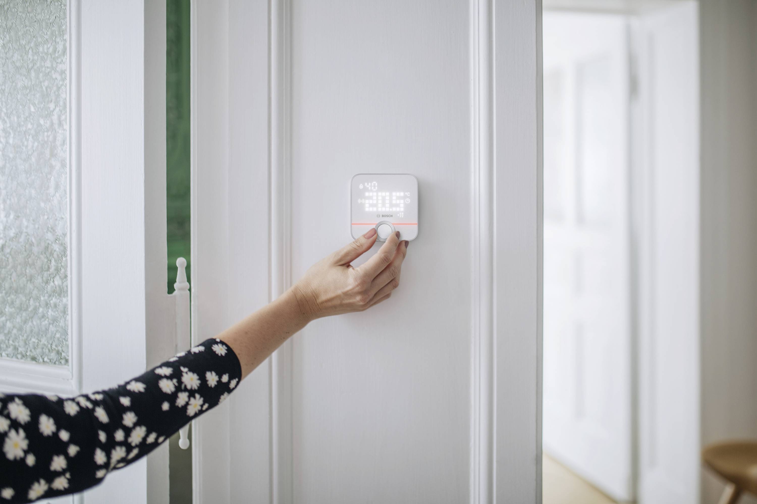 Eine Person stellt ein Thermostat an einer Wand ein, das 20,5 Grad anzeigt. Der Raum im Hintergrund ist hell und offen.