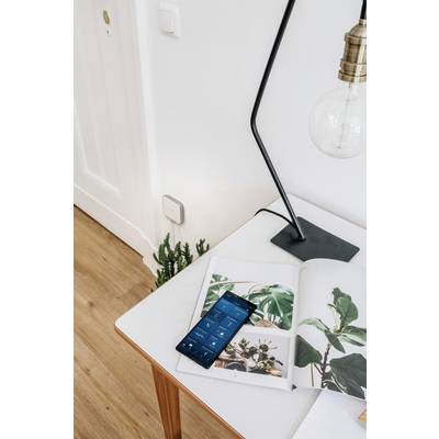 Ein Smartphone liegt auf einem Tisch neben einer geöffneten Zeitschrift mit Pflanzenbildern und einer Lampe.