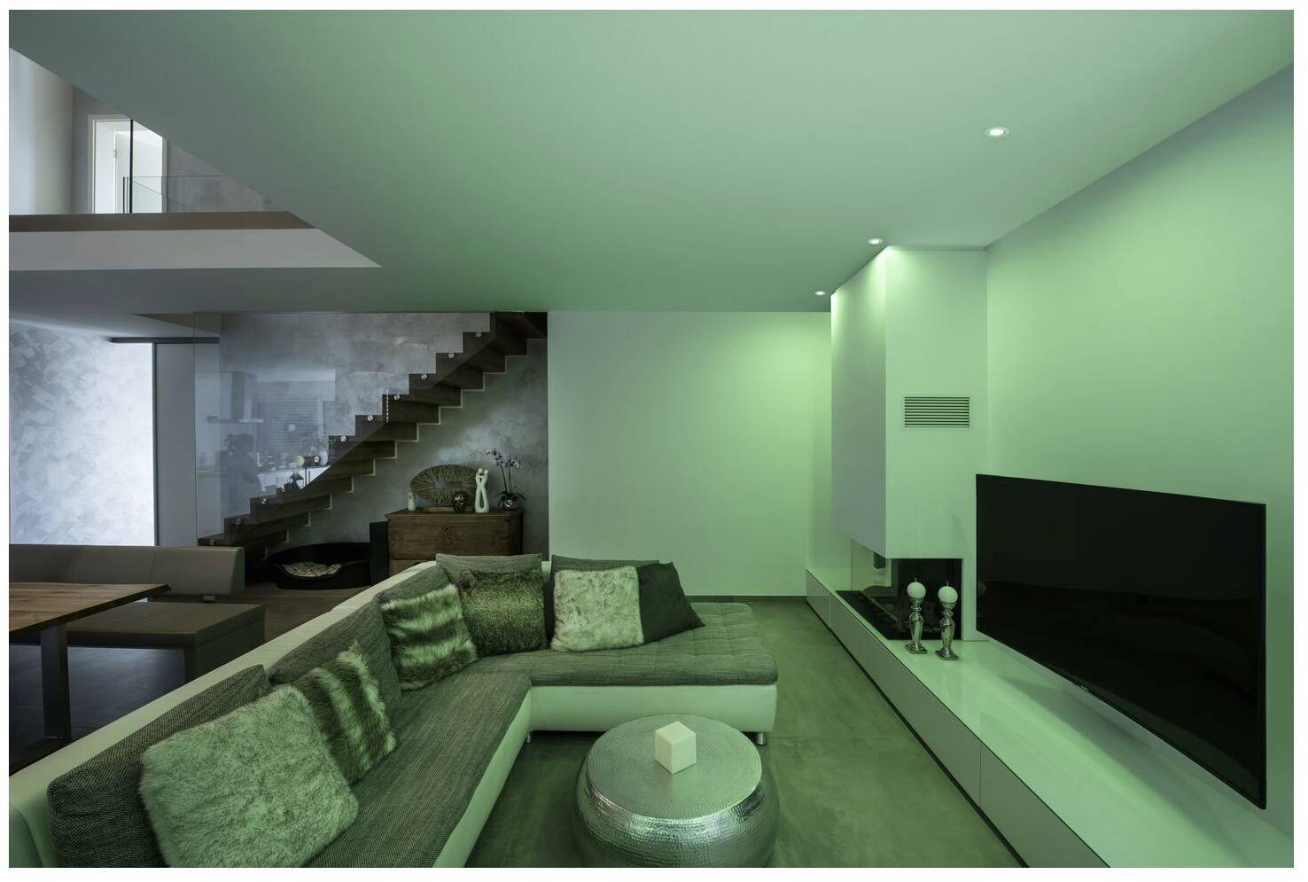 Modernes Wohnzimmer mit grauem Sofa, silbernem Tisch und großem Fernseher. Treppe im Hintergrund, grüne Beleuchtung dominiert den Raum.