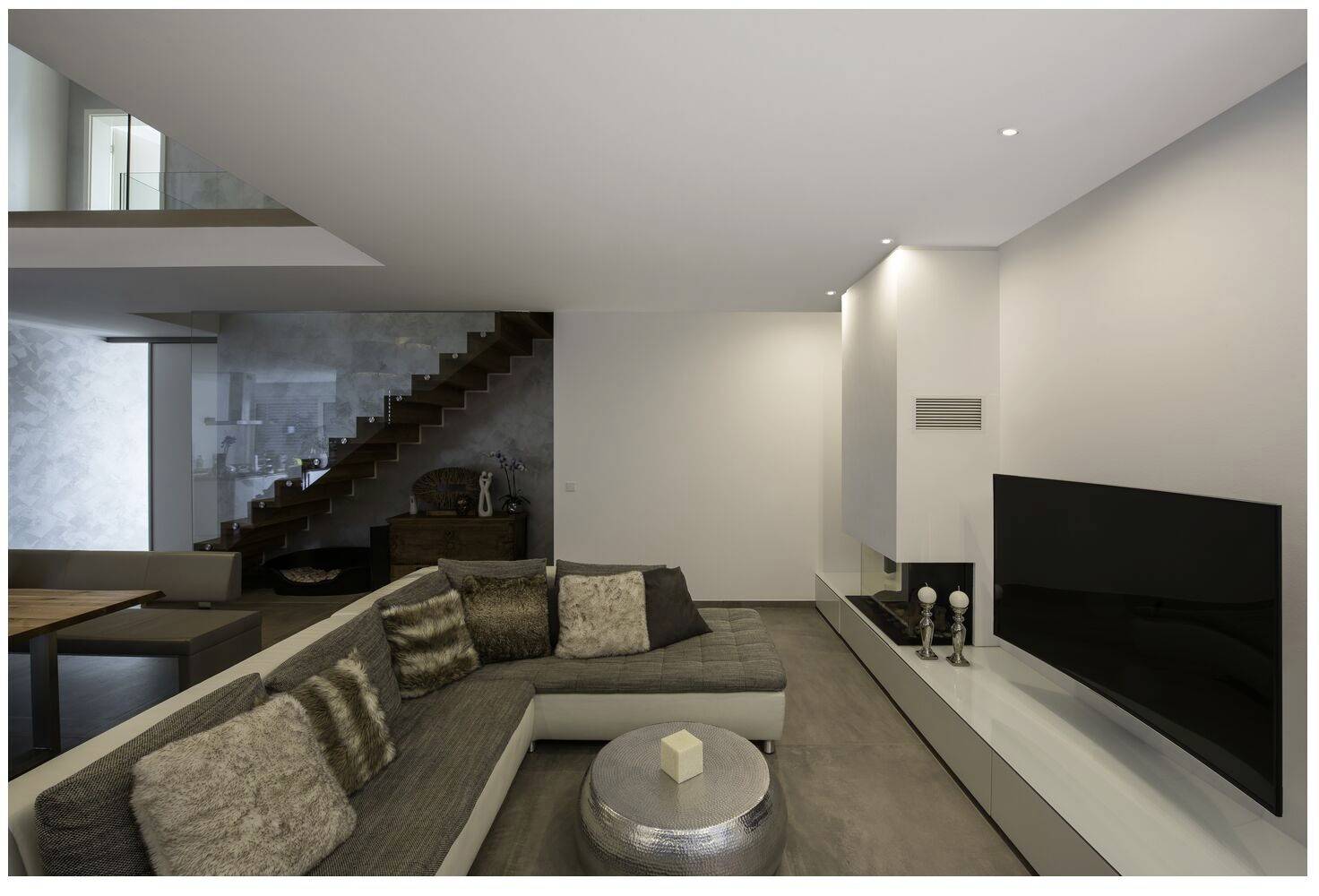 Moderne Wohnzimmer mit grauem Ecksofa, rundem Couchtisch und Flachbildfernseher an der Wand. Offene Treppe führt nach oben.