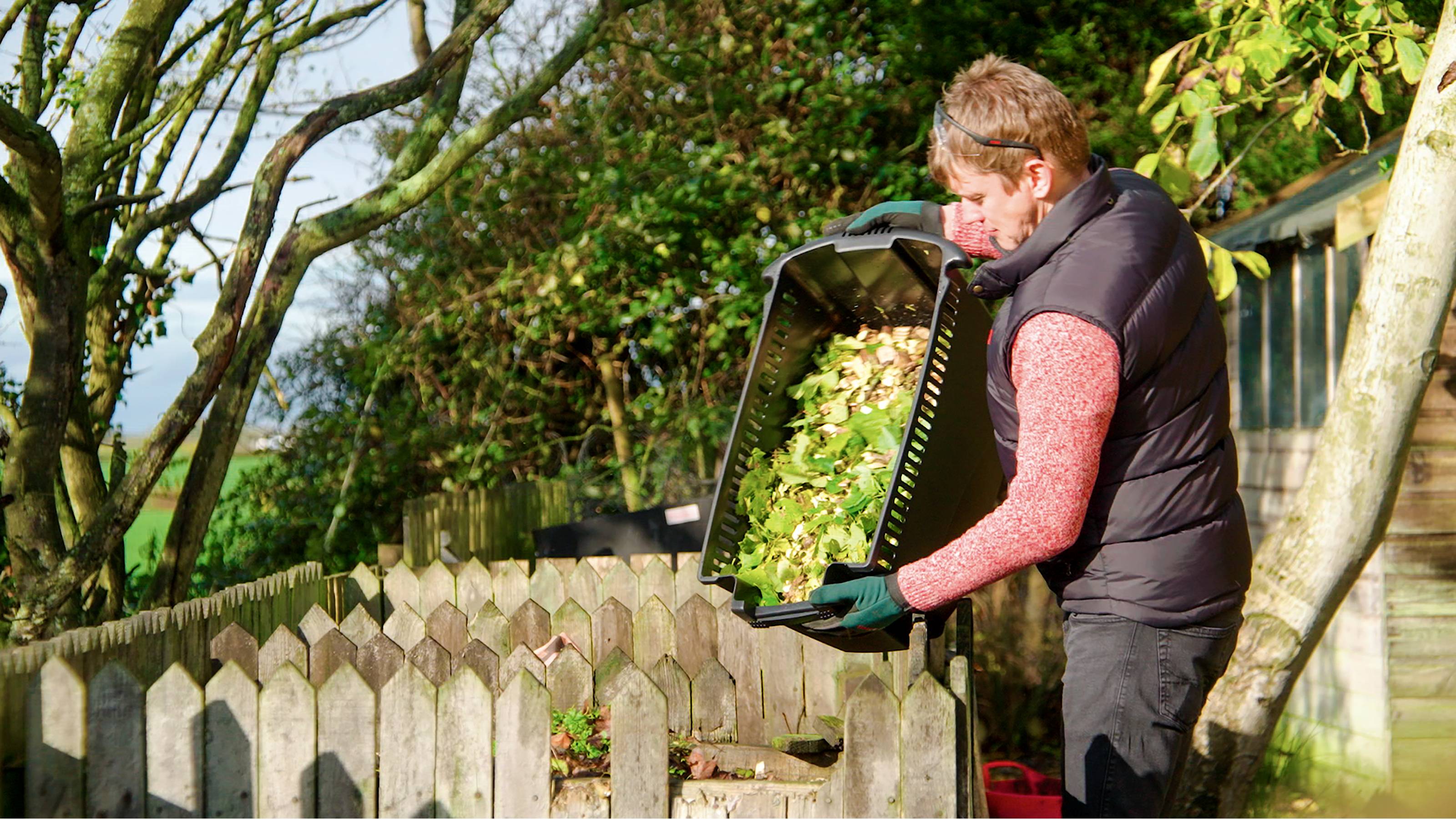 Eine Person leert einen Behälter mit Gartenabfällen in einen Komposter in einem Garten mit Holzzaun und Bäumen im Hintergrund.