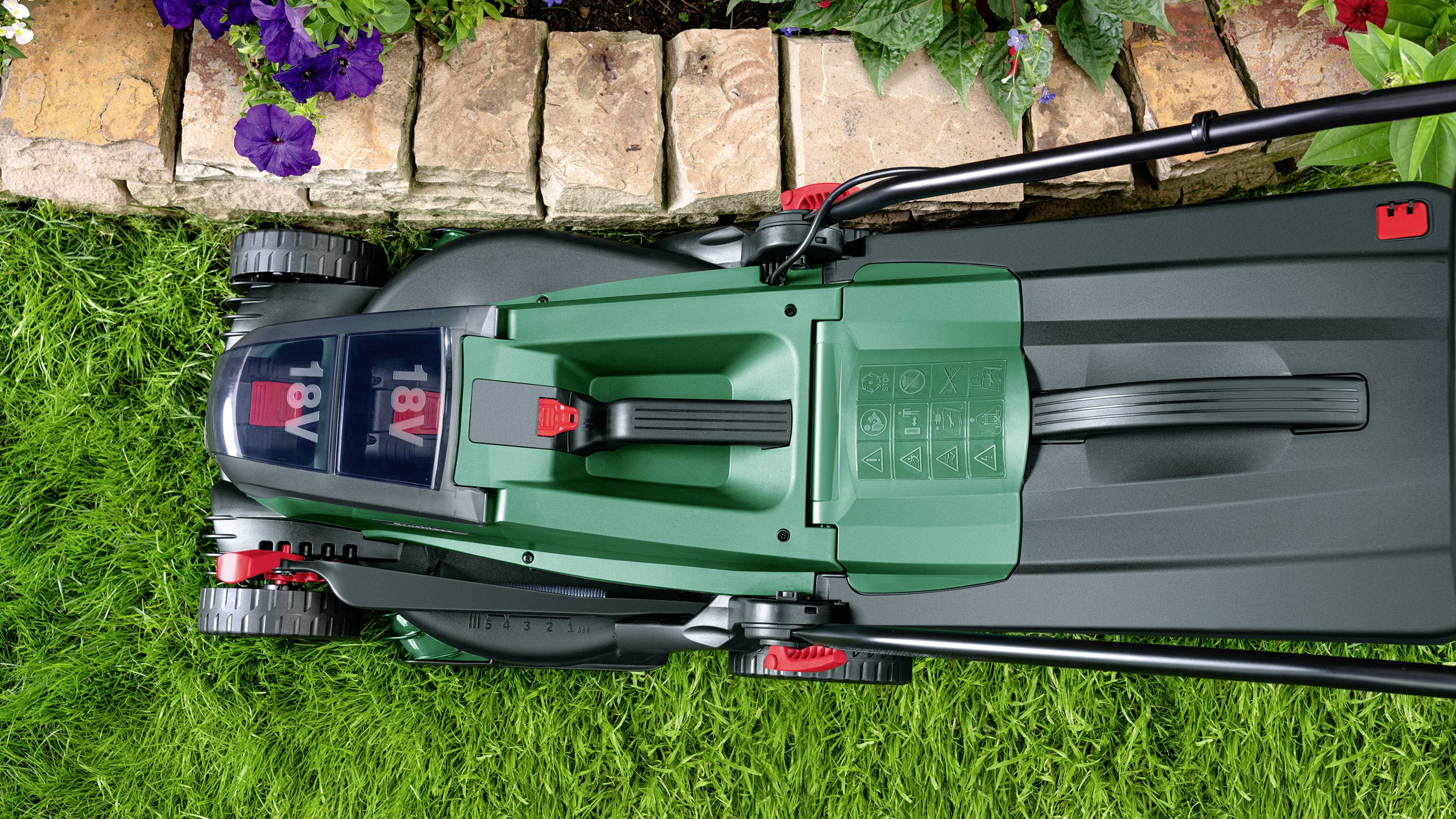 Elektrischer Rasenmäher mäht Rasen entlang einer Blumenbeetkante. Ansicht von oben zeigt grünen und schwarzen Mäher mit Batterieanzeige.