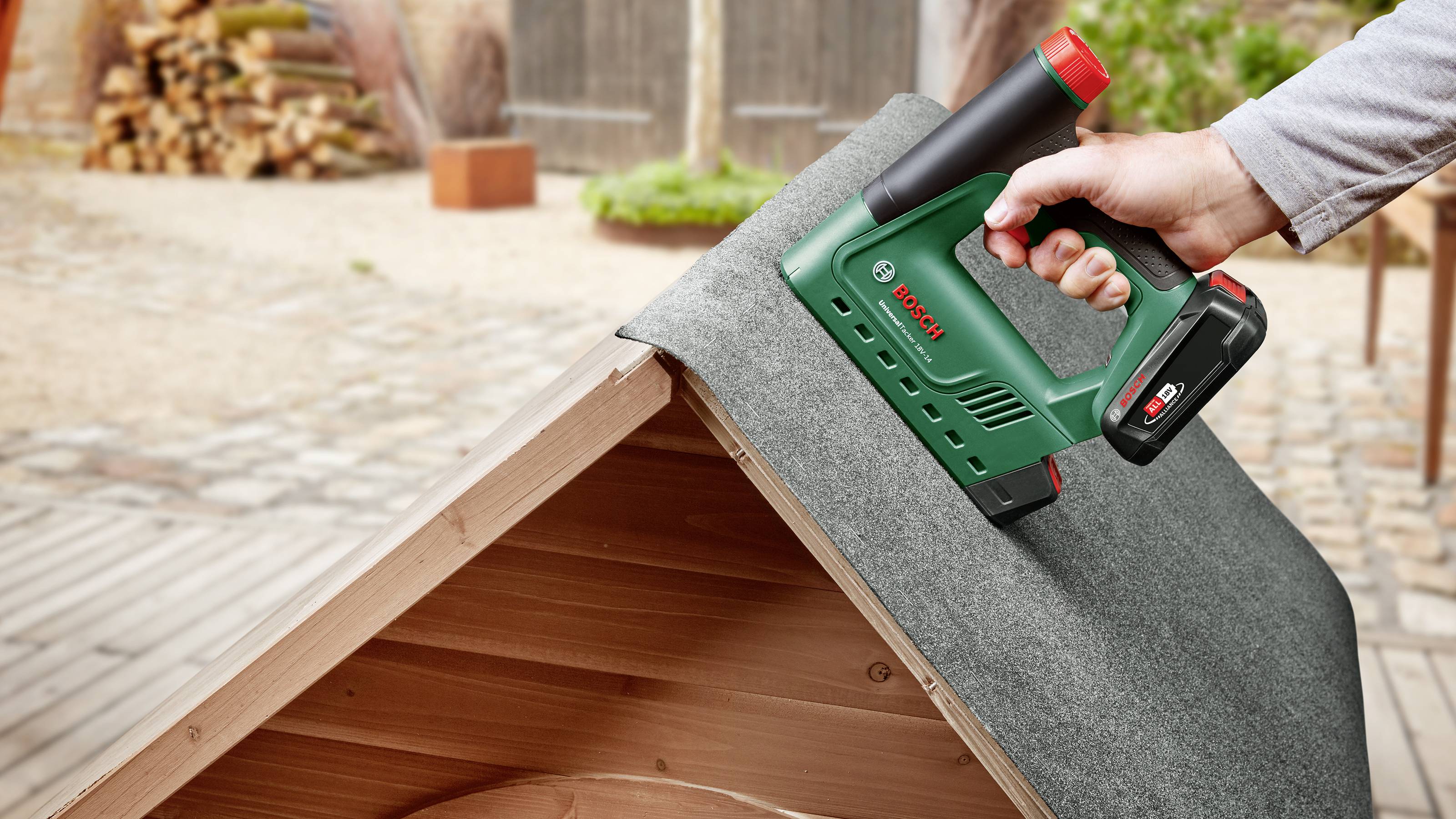 Eine Hand benutzt einen grünen Bosch Elektrotacker, um Dachpappe auf einem kleinen Holzhaus zu befestigen. Im Hintergrund Holzhaufen.