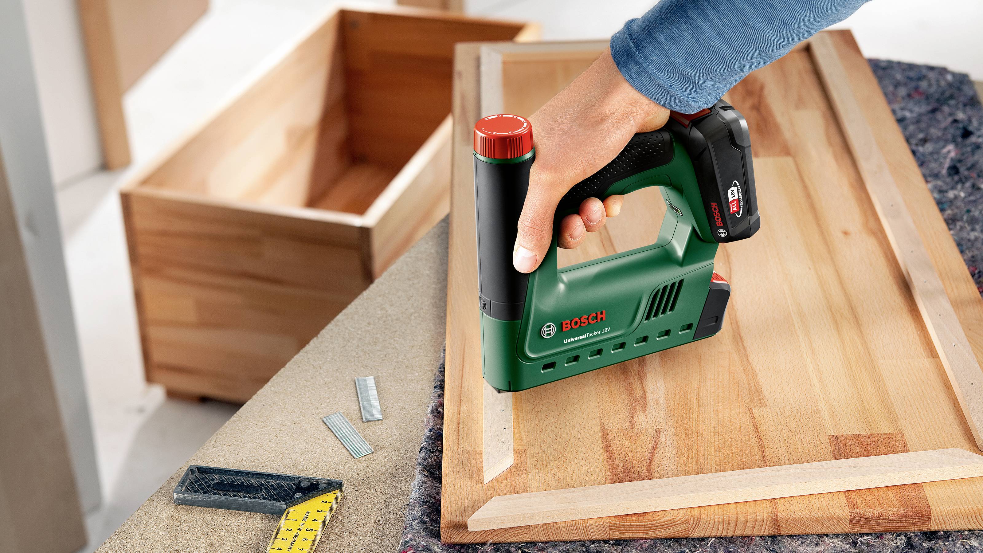 Eine Person verwendet einen grünen Elektrotacker, um Holzstücke auf einem Holzbrett zu befestigen. Im Hintergrund sind Holzkisten zu sehen.