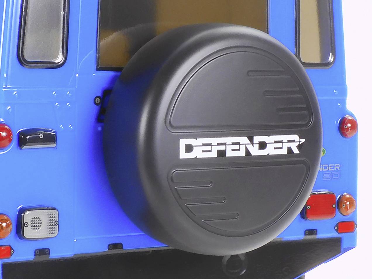 Ein blauer Geländewagen mit einem Reserverad, auf dem 'DEFENDER' steht, montiert am Heck. Fahrzeug hat rechteckige Rücklichter.