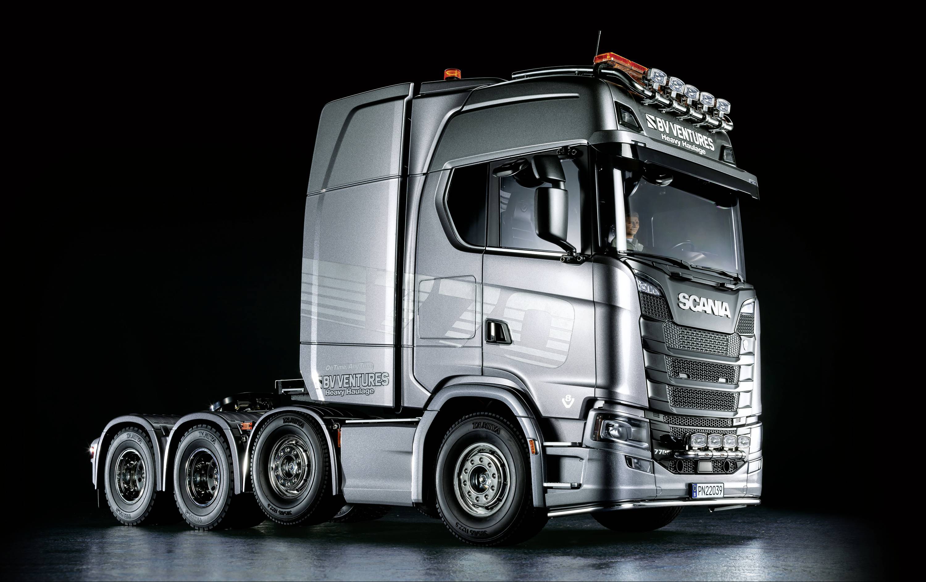 'Ein silberner LKW mit fünf Achsen, markante Beleuchtung auf dem Dach und der Beschriftung 'Scania V8' auf der Seite, vor schwarzem Hintergrund.'